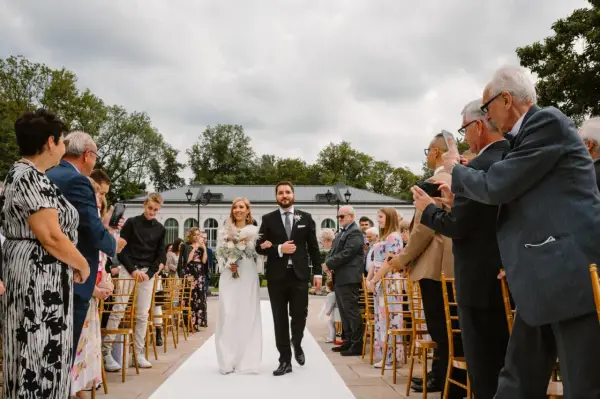 Para młoda idzie alejką, goście robią zdjęcia. Formalności po ślubie w pełni.