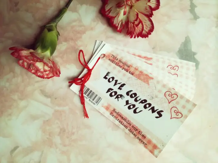 Papierowa rocznica uczczona kuponami miłości. Dwa kwiaty goździka i stosik kuponów "Love Coupons for You" na różowym tle.